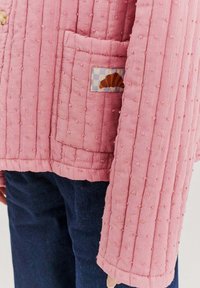 Prošívaná růžová bunda s texturovanou tkaninou, která má přední kapsu a barevnou nášivku croissantu na kostkované ploše.