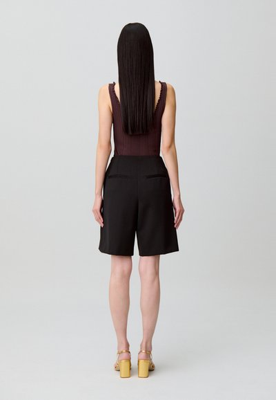 Body de color marrón con textura acanalada y tirantes con volantes, combinado con pantalones cortos negros de corte entallado con bolsillos laterales, acompañado de sandalias amarillas de tacón.