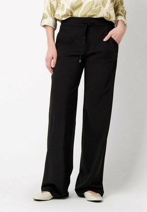 Woman wearing black wide-leg pants with drawstring waist, floral long-sleeve shirt tucked in, and white flat shoes, one hand in pocket.