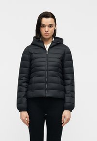 Γυναίκα που φοράει μαύρο καπιτονέ μπουφάν με κουκούλα, κλειστή με φερμουάρ, στέκεται μπροστά σε απλό λευκό φόντο.