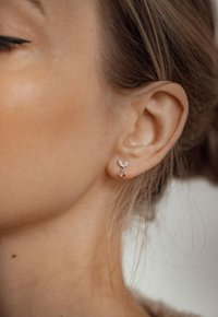 Pendientes de plata en forma de alas de mariposa, con pequeñas piedras preciosas claras, llevados en un primer plano de una oreja. Piel suave y cabello natural visibles.