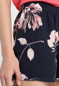 Woman's hand resting beside navy blue shorts with pink and white floral print, paired with a coral pink top.