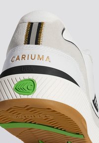 Sneaker bianco e grigio con tomaia textured, caratterizzato da una striscia nera, logo "CARIUMA" dorato e dettaglio verde sulla suola.