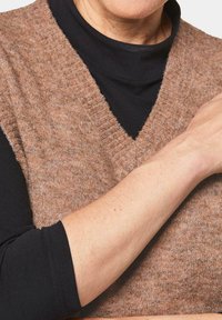 Mouwloze trui van een wolmix in lichtbruin met een diepe V-hals, gedragen over een zwart langemouwen top. Zachte textuur met ribbelaccenten.