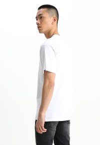 Weißes Baumwoll-T-Shirt mit kurzen Ärmeln, Rundhalsausschnitt und minimalistischem Design, getragen mit dunklen Jeans. Zeigt ein kleines Logo auf dem Ärmel.