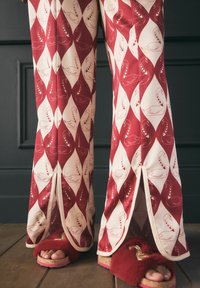 Gemusterte weit geschnittene Hosen zeigen rote und cremefarbene Rauten mit Schwan-Designs. Die Hosen haben Schlitzsäume. Texturierte, flauschige rote Hausschuhe sind sichtbar.