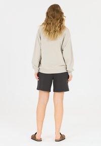 Ljusgrå sweatshirt med lös passform och ribbad fåll, kombinerad med mörkgrå shorts. Håret i lösa vågor, stående framför en enfärgad bakgrund.