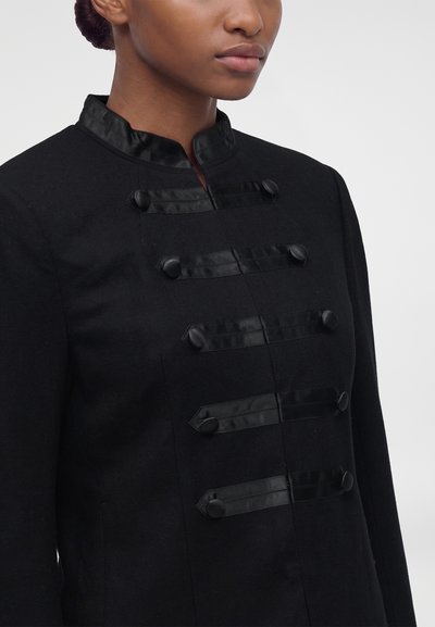Femme portant une veste noire avec un col montant et une finition en satin avec cinq sangles horizontales décoratives à boutons.
