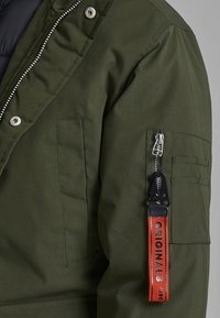 Green parka jacket featuring a zippered front, large side pocket, and an orange fabric tag labeled "ORIGINALS" on the sleeve.