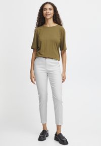 Une femme aux cheveux bouclés sourit en portant une chemise verte olive, un pantalon gris clair et des chaussures noires, se tenant contre un fond uni.