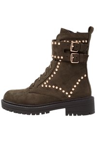 Bottines en daim vert olive avec des clous décoratifs en or rose, des sangles à deux boucles et une semelle en caoutchouc noir épaisse. Design à lacets à l'avant.