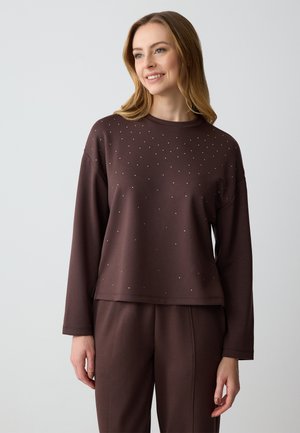 Maglietta marrone a maniche lunghe con vestibilità comoda, caratterizzata da piccoli dettagli metallici sul davanti. Texture arricciata lungo i bordi.