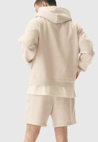 Haut à capuche beige avec une grande capuche, des manches amples et un ourlet superposé, associé à un short assorti en tissu doux et texturé.