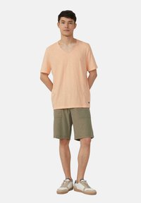 Jasnopomarańczowa koszulka z dekoltem w serek w luźnym kroju, sparowana z oliwkowozielonymi szortami cargo. Model nosi beżowe trampki z brązowymi akcentami.