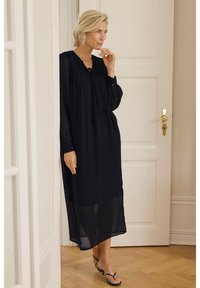 Black, long-sleeved dress in lightweight fabric with a pleated design, features a tie neckline and split sides, worn with minimalistic sandals.