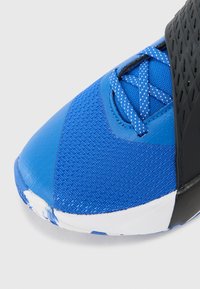 Chaussure de sport bleue avec une tige en tissu texturé, un nez en synthétique lisse, des accents noirs en caoutchouc contrastants, et des lacets bleus avec des éclats blancs.
