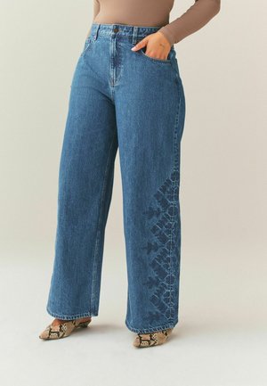 Frau trägt hoch taillierte, weit geschnittene blaue Jeans mit dunkler Blumenstickerei am rechten Bein, kombiniert mit braunen spitz zulaufenden Schuhen mit Schlangenmuster.