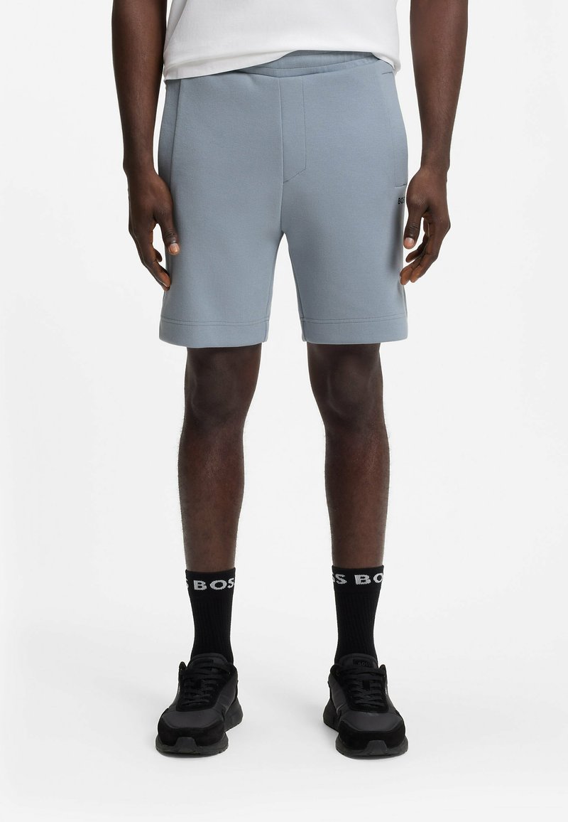 Homme portant un short bleu clair, des chaussettes noires avec texte blanc et des baskets noires, debout sur un fond blanc.