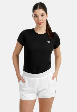 Femme aux cheveux bruns attachés en queue de cheval, portant un t-shirt noir à manches courtes et un short blanc, les mains dans les poches, devant un fond uni.