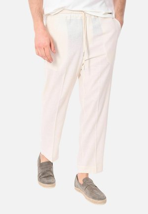 Uomo che indossa pantaloni bianchi con coulisse e scarpe slip-on in camoscio grigio, in piedi con le mani nelle tasche su uno sfondo semplice.