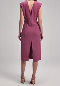 Robe ajustée longueur genou en rose poudré avec manches courtes, dos en V profond et fente arrière. Complétée par des talons dorés à brides.