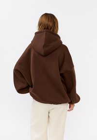 Oversized bruine hoodie met een grote capuchon en afgeknipte schouders, gemaakt van zachte stof. Gedomineerd met crèmekleurige broek, die een relaxte pasvorm laat zien.