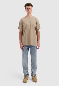 Jonge man die staat te poseren in een beige "PURE PATH" T-shirt, lichtblauwe jeans en beige veterschoenen tegen een effen lichtgrijze achtergrond.