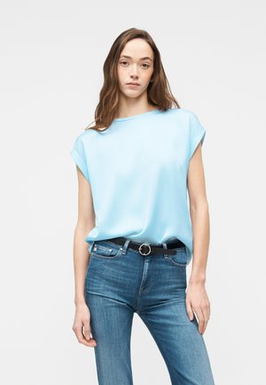 Młoda kobieta ubrana w jasnoniebieski, bezrękawnik włożony w niebieskie dżinsy z średnim stanem i czarnym paskiem, stojąca na jednolitym tle.