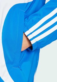 Blue athletic jacket with white stripes on sleeve, featuring elastic cuffs and a side pocket. Made from smooth, lightweight fabric.