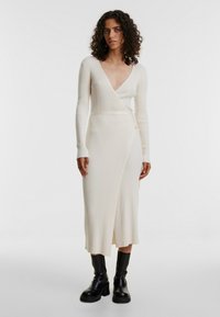 Geripptes, cremefarbenes Wickelkleid mit langen Ärmeln, tiefem V-Ausschnitt und gebundenem Taillengürtel, kombiniert mit schwarzen Knöchelschuhen. Glatte Textur und figurbetonte Form.