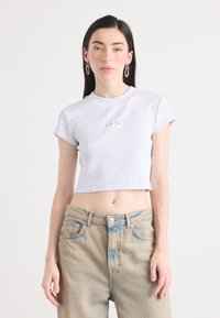 Model in een cropped lila t-shirt met het 'Calvin Klein' logo, draagt lichtblauwe high-waisted jeans, tegen een simpele witte achtergrond.