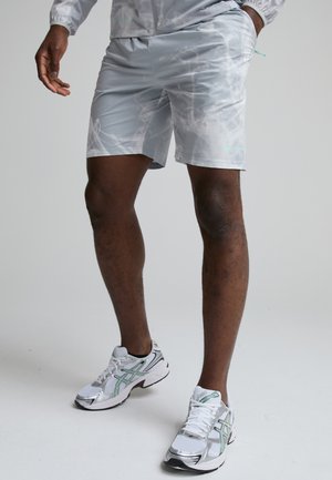 Homme portant un short gris clair à motifs, une main dans la poche, debout en chaussures de course blanches et argentées sur fond uni.