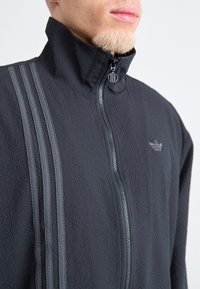 Giacca in pile nera con zip, dotata di colletto alto, realizzata in tessuto testurizzato, con tre strisce grigie lungo il lato sinistro e un piccolo logo sul petto.