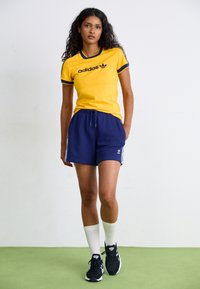 Gelbes Adidas T-Shirt mit navyblauen Details und Logo; navyblaue Shorts mit weißen Seitenstreifen; schwarze Sportschuhe; weiße Socken. Grüner Bodenbelag.