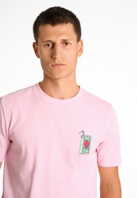 Ljust rosa bomulls t-shirt med en grafisk fickdesign som har ett dryckeförpackning och ett rött hjärtmotiv. Kort ärm och rund halsringning.