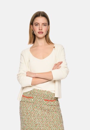 Frau mit verschränkten Armen, trägt ein weißes Oberteil und eine Strickjacke sowie einen Blumenrock mit orangefarbenen Akzenten, blickt mit neutralem Gesichtsausdruck nach vorne.