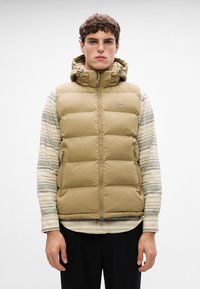 Gilet imbottito color khaki con cappuccio, dotato di zip frontale e due tasche laterali. Indossato sopra una camicia a righe a maniche lunghe e mostrato in un ambiente neutro.