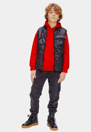 Schwarze gesteppte Weste über einem roten Kapuzenpullover, kombiniert mit dunklen grauen Cargo-Hosen und schwarzen High-Top-Sneakern mit einer beigefarbenen Sohle.