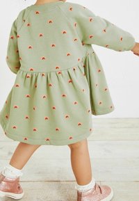 Abito in cotone verde con maniche lunghe, gonna arricciata, caratterizzato da piccoli arcobaleni ricamati in arancione e rosso. Indossato con scarpe luccicanti.