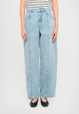 Person som har på seg høymidjede, lyseblå vide jeans med synlige frontseam, en stripete skjorte stukket inn, og svarte firkantede sko.