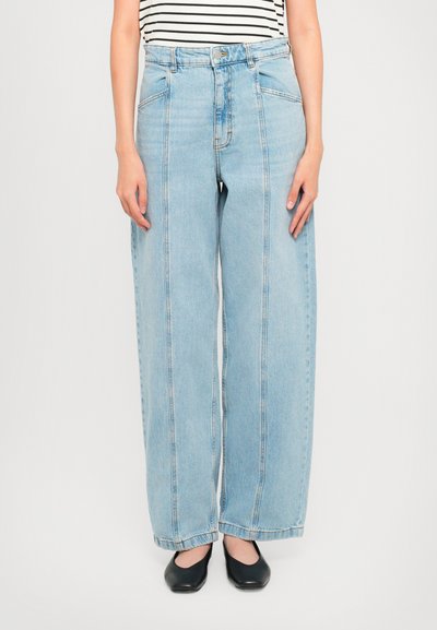 Person iført højtaljede, lyseblå, brede jeans med synlige forreste sømme, en stribet skjorte stoppet ind, og sorte firkantede sko.