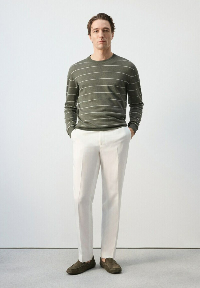 Homme debout, mains dans les poches, portant un pull rayé vert olive, un pantalon blanc et des mocassins marron, sur un fond uni.