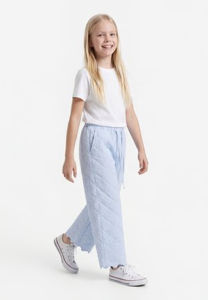 Ung pige med langt blondt hår, der går, iført en hvid t-shirt, lyseblå teksturerede bukser og hvide sneakers.