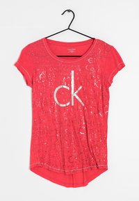 Camiseta roja de manga corta, con un gran logo en plata de "ck" y un diseño de texto repetido. Tejido suave con un escote redondeado y un dobladillo asimétrico.