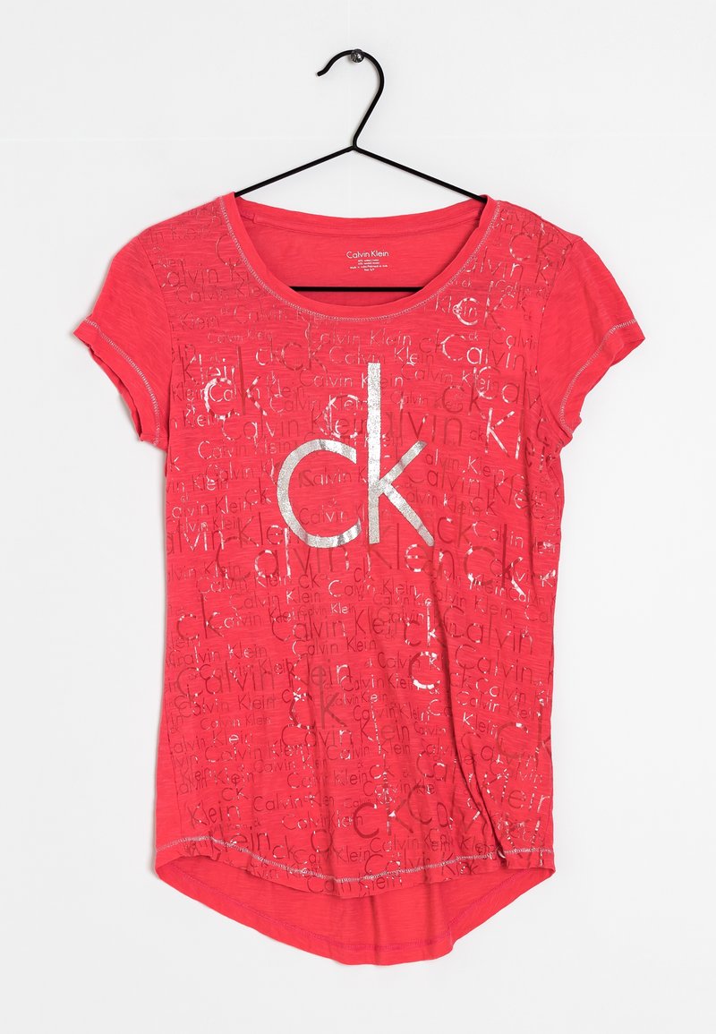 Camiseta roja de manga corta, con un gran logo en plata de "ck" y un diseño de texto repetido. Tejido suave con un escote redondeado y un dobladillo asimétrico.