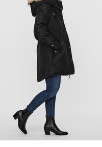 Parka noire isolée avec une capuche en fausse fourrure, fermeture éclair et boutons-pression, et poches latérales ; associée �à un jean bleu foncé et des bottines noires à talons.