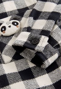 Schwarz-weiß kariertes Flanellhemd mit weicher Textur, versehen mit einem Pandapatch und einem schwarzen Knopf an der Manschette.
