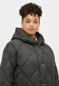 Podstavljena jakna s kapuljačom u tamnozelenoj boji, s prednjim kopčanjem na dugmad i dijamantnim uzorkom. Mekana, glatka tkanina.