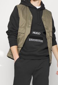 Mouwloos olijfgroene vest over een zwarte hoodie, met een logo en zakdetails. Zwarte joggingbroek maakt de outfit compleet. Getextureerde stof.