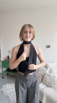 Svart ärmlös topp och grå vida byxor. En mörk randig slips lindas runt halsen. Ljus bakgrund med mjuka inredningsdetaljer.